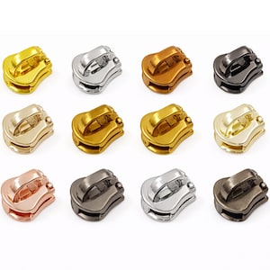 May include: A collection of twelve metal zipper pulls in various metallic finishes, including gold, silver, bronze, and black. Each pull has a curved design with a raised handle for easy gripping. The pulls are arranged in a grid pattern.