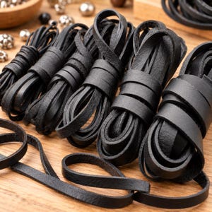 May include: Several coils of flat black leather cord arranged in a row. The leather strips have a matte texture and are wound into tight, circular bundles. A few loose strands of the black leather cord extend outward from the main coils.