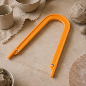 May include: A U-shaped orange clay cutter tool featuring a thin metal wire stretched between two plastic arms, secured by two silver-coloured screws at the base. The tool is designed for slicing through clay with a simple, functional frame.