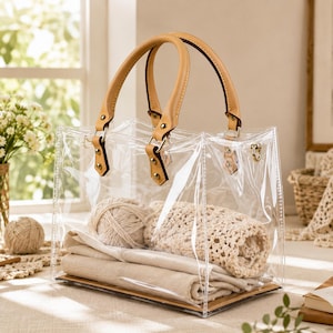 May include: A clear, square-shaped tote bag with two tan leather handles attached by gold-toned metal hardware. The bag contains a ball of beige yarn, a folded piece of light-colored fabric, and a textured, cream-colored crocheted textile piece.