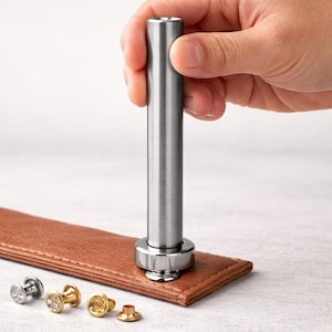 May include: A metal rivet setter tool is used to set a silver rivet on a brown leather strap. Several other rivets in silver and gold are visible. The tool is held by a hand.