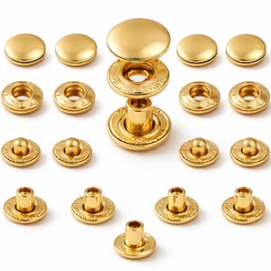 Puede incluir: Una colección de cierres de presión dorados, que incluyen componentes de tapa, casquillo y espárrago. Los cierres están dispuestos sobre un fondo blanco, mostrando su diseño y construcción. La ferretería metálica es probablemente para proyectos de ropa o marroquinería.