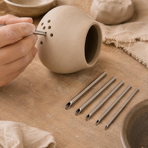 May include: A round, light beige, unglazed ceramic vessel with a wide opening and a cluster of small circular perforations on its side. Five thin, silver-coloured metal tools with angled, hollow tips are arranged in a row on the surface.