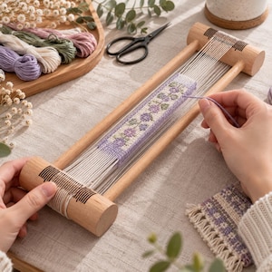 Op de afbeelding: Een houten kralenweefgetouw met een rechthoekig frame en twee cilindrische uiteinden met gleuven. Witte scheringdraden zijn over het frame gespannen en houden een gedeeltelijk geweven band met een paars en groen bloemenpatroon vast.