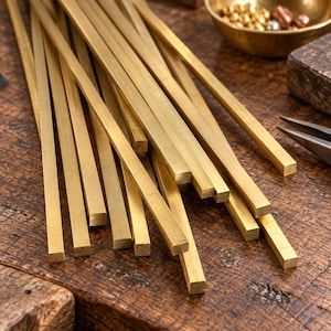 Könnte beinhalten: Eine Ansammlung langer, dünner Messingstäbe mit quadratischem Profil und metallisch goldener Oberfläche. Die Stäbe sind in einem losen, überlappenden Bündel angeordnet und zeigen ihre geraden, gleichmäßigen Kanten und glatten Oberflächen.