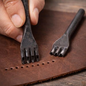 May include: Close-up of leatherworking tools. A hand holds a metal stitching chisel over a piece of brown leather. The chisel has multiple prongs, creating evenly spaced holes. Another chisel and a row of holes are also visible.