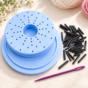 Peut inclure: Un outil de tissage circulaire en plastique bleu clair avec un trou central et des trous numérotés disposés en cercles concentriques, accompagné d'un tas de petits piquets en plastique noir et d'une aiguille à laine violette.