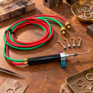 Op de afbeelding: Een soldeerbrander voor sieraden met een zwarte handgreep, zilveren en blauwe instelknoppen en een koperen punt. Het bevat een paar rood en groen gevlochten slangen met messing fittingen en vier verwisselbare koperen tips in diverse hoeken.