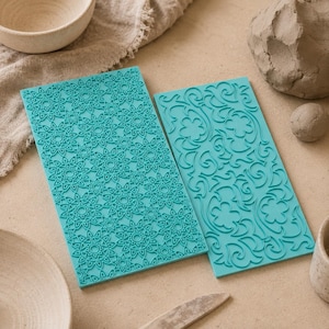 Puede incluir: Dos alfombrillas de textura rectangulares de silicona turquesa para arcilla. Una presenta un patrón geométrico de mandala repetitivo, mientras que la otra muestra un diseño intrincado de enredaderas florales con pétalos en relieve.