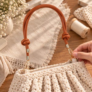Pode incluir: Uma bolsa de crochê na cor creme com um design plissado e texturizado. A bolsa inclui uma alça arredondada e lisa na cor castanho-claro com nós decorativos em cada lado, fixada à bolsa com fechos de mosquetão de metal dourado polido.