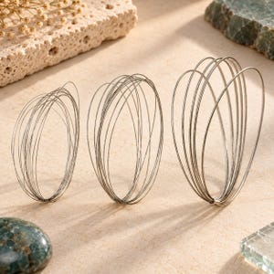 May include: Three sets of thin, silver-colored metal wire loops arranged in oval shapes of varying sizes. Each set consists of multiple concentric wire strands that overlap to create a hollow, cage-like structure with a metallic finish.