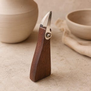 Peut inclure: Un outil de poterie manuel doté d'un manche en bois brun foncé et d'une lame métallique argentée incurvée, fixée par une vis cruciforme. L'outil se tient debout sur sa base plate, mettant en valeur une finition en bois lisse et polie.