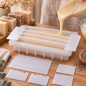 Peut inclure: Un moule à savon rectangulaire en silicone blanc avec plusieurs séparateurs internes, en cours de remplissage avec un liquide beige crémeux. Le kit comprend divers inserts diviseurs en plastique blanc, plats ou crantés, posés sur une surface en bois.