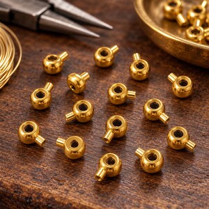 Könnte beinhalten: Eine Ansammlung kleiner, glänzend goldfarbener Metallperlen, die jeweils aus einem kugelförmigen Körper mit einer kurzen, zylindrischen Öse bestehen. Diese Metallkomponenten sind auf einer dunklen Holzoberfläche neben einer Zange angeordnet.