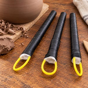 May include: Three pottery trimming tools with long, cylindrical black handles wrapped in tape. Each tool features a white base section and a bright yellow wire loop at the end, designed for shaping and carving clay on a pottery wheel.