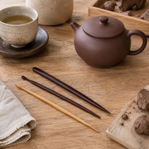 Può includere: Una teiera in argilla marrone scuro con coperchio, una tazza da tè in ceramica chiara su un piattino in legno scuro e tre sottili strumenti per scolpire in legno in varie tonalità di marrone chiaro e scuro su una superficie in legno.