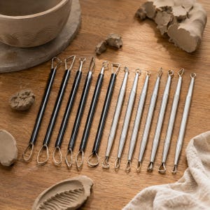 Könnte beinhalten: Ein Set aus dreizehn Töpfer-Modellierwerkzeugen mit dünnen Metalldrahtschlaufen an beiden Enden. Sechs Werkzeuge haben schwarze zylindrische Griffe, während sieben Werkzeuge silberfarbene Metallgriffe aufweisen, angeordnet in einer Reihe.