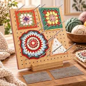 Könnte beinhalten: Ein quadratisches Spannbrett aus Holz mit einem Lochraster und einem Standfuß, in das mehrere Metallstifte eingesetzt sind, um ein Granny Square, ein florales Granny Square, ein rundes Deckchen und ein dreieckiges Häkelstück zu fixieren.