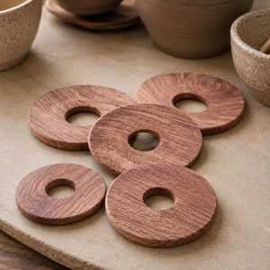 May include: A set of five circular wooden coasters, each featuring a central hole. The coasters are crafted from brown wood with a visible natural grain pattern, arranged in a cluster on a flat, light-coloured surface with a textured appearance.