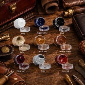 Op de afbeelding: Negen kleine ronde potjes met leerkleur of crème in diverse kleuren: wit, saffierblauw, zwart, beige, lichtbruin, middenbruin, wijnrood, grijs en donkerrood. Elk potje heeft een bijpassend deksel en een gedrukt label met de kleurnaam.