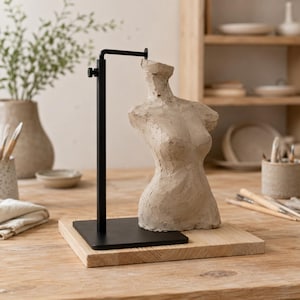 Può includere: Una scultura di torso in argilla beige montata su un espositore in metallo nero con un braccio orizzontale regolabile. Il supporto è fissato a una base quadrata in legno, fornendo una piattaforma stabile per la figura dalla forma testurizzata.