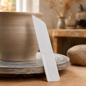 May include: A white plastic pottery rib tool with a flat, angled edge and a rectangular handle indentation, positioned upright next to a light brown clay pot resting on a circular wooden pottery wheel base in a studio setting.