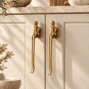 May include: Two gold-colored cabinet handles shaped like stylized figures sitting with their hands resting on their faces. The figures have elongated legs that hang down the front of the cream-colored cabinet doors, featuring a smooth metallic finish.