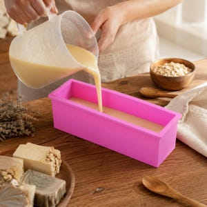 Puede incluir: Un molde rectangular de silicona rosa siendo llenado con un líquido beige claro vertido desde una jarra medidora de plástico transparente. Cerca hay varias barras de jabón terminadas en un plato de madera y un cuenco de madera con avena.