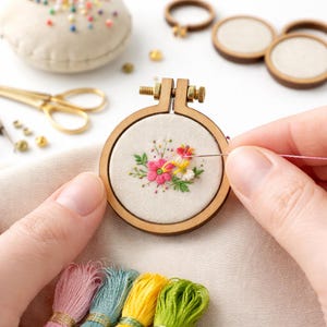 Op de afbeelding: Een kleine houten borduurring met een bloemenontwerp in roze, geel en wit. Er wordt geborduurd met een naald en roze draad. Andere borduurbenodigdheden, waaronder een schaar en draad, zijn zichtbaar.