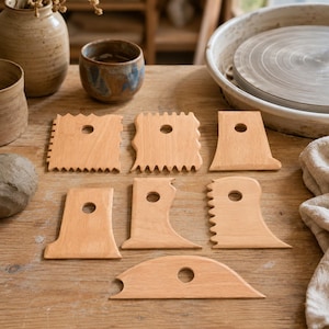 Può includere: Un set di sette strumenti per ceramica in legno chiaro, ciascuno dotato di un foro circolare centrale. Questi utensili presentano vari bordi, inclusi profili seghettati, dritti e curvi, progettati per modellare e levigare l'argilla.