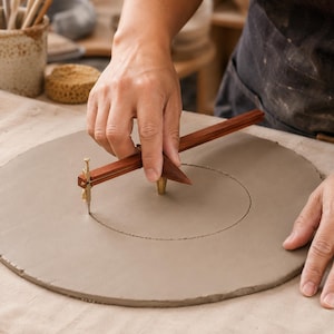 May include: A wooden pottery compass tool with brass hardware and a sharp metal point, used to scribe a precise circle into a flat, circular slab of grey clay. The tool features a long wooden arm and a central pivot point for consistent rotation.