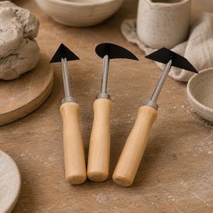 May include: Three pottery trimming tools with light-coloured wooden handles and silver metal shafts. Each tool features a distinct black metal blade: one triangular, one curved, and one wide trapezoidal shape, designed for clay sculpting.