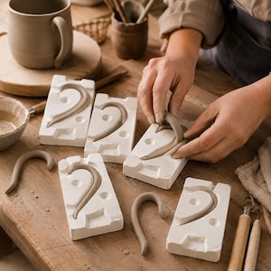 May include: A set of rectangular white plaster molds for ceramic handles, each featuring a curved indentation. One mold is being used to shape a smooth, light gray clay handle. Several finished clay handles lie nearby on a wooden surface.