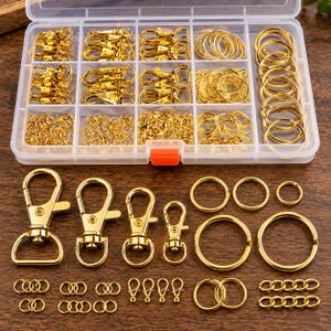 Könnte beinhalten: Ein durchsichtiger Kunststoff-Aufbewahrungsbehälter gefüllt mit goldfarbenen Metall-Schmuckzubehörteilen, darunter verschiedene Größen von Karabinerverschlüssen, Spaltringen, Biegeringen und kurzen Kettengliedern, die auch lose davor liegen.