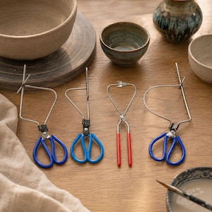 May include: Four metal pottery tongs with scissor-style handles. Three tongs feature blue rubber-coated handles and one features red rubber-coated handles. The metal arms are shaped into different configurations for gripping ceramic vessels.