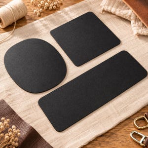 May include: Three matte black coasters with smooth surfaces, consisting of one circular coaster, one square coaster with rounded corners, and one rectangular coaster with rounded corners, arranged on a light beige fabric surface.