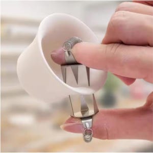 May include: A silver-colored metal finger guard featuring a curved shield and a coiled spring ring. The device has two pointed prongs extending from the base of the shield, designed to protect the fingertip during cutting or slicing tasks.