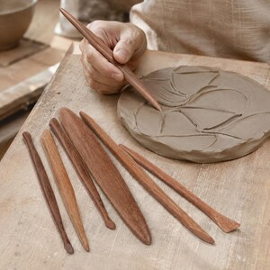May include: A set of seven smooth, brown wooden pottery sculpting tools of varying shapes and sizes, including pointed, flat, and rounded ends, arranged on a work surface next to a circular piece of grey clay being carved with a pointed tool.