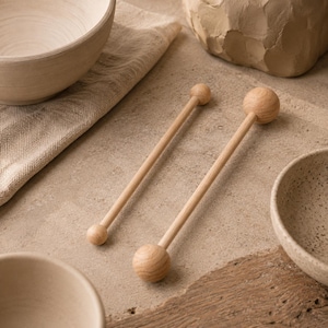 May include: Two light-coloured wooden stirrers, each featuring a long, slender cylindrical handle capped with a small spherical knob at one end and a larger spherical knob at the opposite end, showcasing a natural wood grain texture throughout.