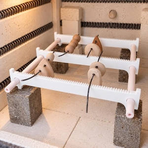 May include: A white ceramic kiln shelf rack with notched edges and light pink cylindrical rods, supporting several small, unglazed clay beads and pendants threaded onto dark metal wires, all resting on four rectangular refractory bricks.