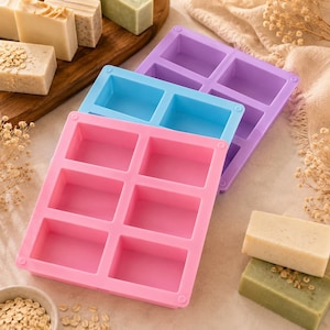 Peut inclure: Trois moules rectangulaires en silicone pour la fabrication de savon, empilés en quinconce. Le moule supérieur est violet, celui du milieu est bleu et celui du bas est rose. Chaque moule comporte six cavités rectangulaires pour le savon.