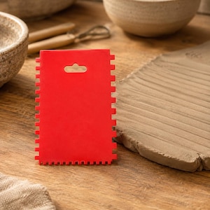 May include: A rectangular red pottery rib tool featuring a serrated edge with teeth along three sides and a small cutout hole at the top center. The tool is constructed from a flexible material and is designed for shaping and texturing clay surfaces.
