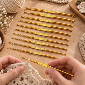 Op de afbeelding: Een set dubbelzijdige goudkleurige metalen haaknaalden, elk voorzien van gegraveerde maataanduidingen zoals 3/0, 4/0, 5/0, 7/0, 7.5/0, 8/0 en 10/0. Eén naald is in gebruik, terwijl de andere parallel op een houten ondergrond liggen.