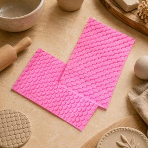 Puede incluir: Una alfombrilla de silicona rectangular de color rosa brillante con un patrón repetitivo en relieve de escamas de pez o vieiras en toda su superficie. La alfombrilla flexible está diseñada para crear impresiones texturizadas en arcilla.
