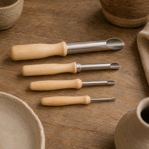 May include: A set of four metal tools with light-coloured wooden handles, arranged in descending order of size. Each tool features a cylindrical metal shaft that terminates in a slanted, open-ended scoop or gouge tip, designed for precision work.