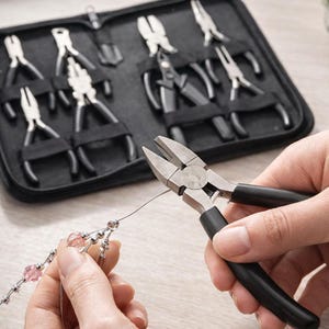 Puede incluir: Un juego de herramientas para hacer joyas, que incluye alicates, exhibido en un estuche negro. Se utiliza un par de alicates con mangos negros para manipular un alambre con cuentas rosas. Las herramientas son para manualidades y joyería.