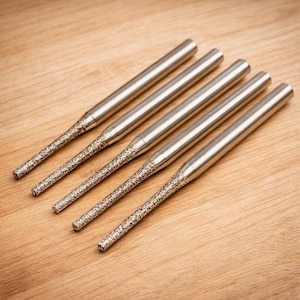 Może przedstawiać: Pięć srebrnych frezów obrotowych z teksturowanymi, ściernymi końcówkami. Frezy ułożone są po przekątnej na jasnobrązowej drewnianej powierzchni, ukazując ich cylindryczny kształt i szczegółową konstrukcję. Narzędzia te przeznaczone są do precyzyjnej pracy.