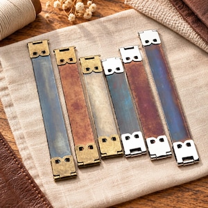 Pode incluir: Seis conectores de alça retangulares de metal com extremidades articuladas, dispostos em uma fileira. Os conectores apresentam acabamentos variados, incluindo latão antigo, cobre e prata polida, cada um com dois pequenos recortes.