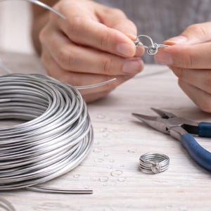 Może przedstawiać: Zbliżenie na dłonie osoby tworzącej srebrny pierścionek z drutu. Widoczna jest również szpula srebrnego drutu i szczypce z niebieskim uchwytem. Gotowy pierścionek znajduje się obok szczypiec. Pierścionek wykonany jest z wielu warstw drutu.