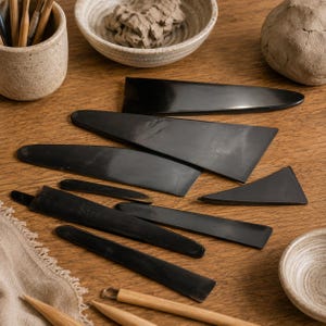Könnte beinhalten: Eine Sammlung schwarzer Töpferrippen und Glättwerkzeuge in verschiedenen Formen und Größen, angeordnet auf einer Holzoberfläche, begleitet von einem Keramikbecher mit Holzwerkzeugen, einer kleinen Schale mit Ton und einem Klumpen Ton.